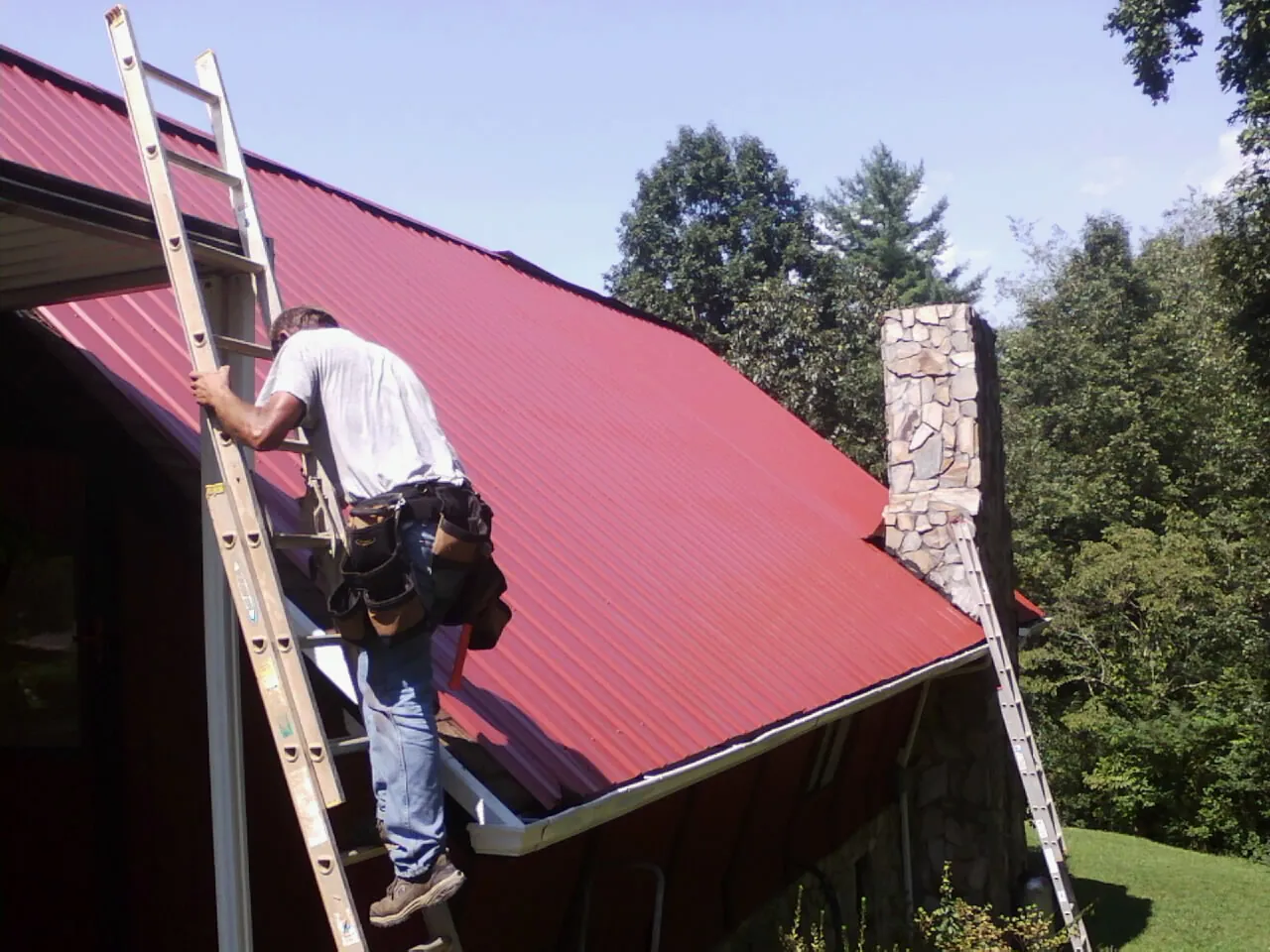Roofer on a ladder beside a red metal roof and stone chimney — Soffit & Fascia Repair by Roofing Brothers in Carrier